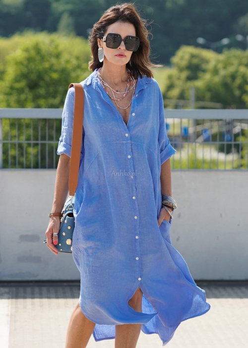 Niebieska lniana sukienka maxi w stylu szmizjerki, zapinana na guziki, o luźnym fasonie oversize, stylizowana z sandałami i torebką na ramię.