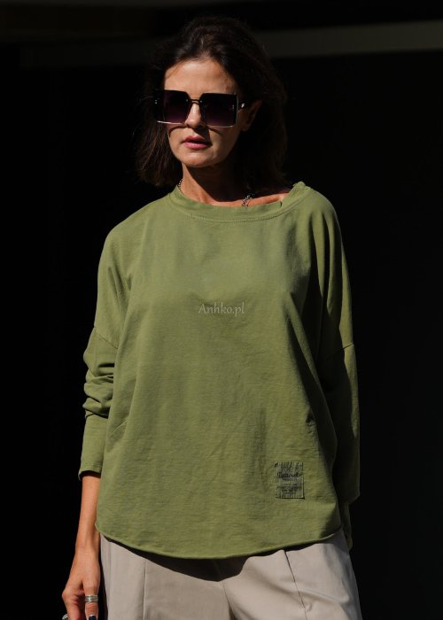 Zielona bluza oversize Nora z obniżoną linią ramion, długimi rękawami i dekoracyjną naszywką przy dole, zestawiona ze spodniami w podobnej tonacji.