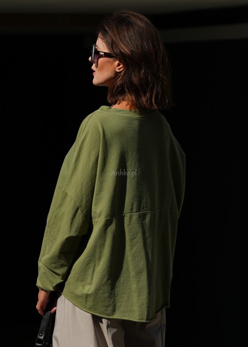 Zielona bluza oversize Nora z obniżoną linią ramion, długimi rękawami i dekoracyjną naszywką przy dole, zestawiona ze spodniami w podobnej tonacji.