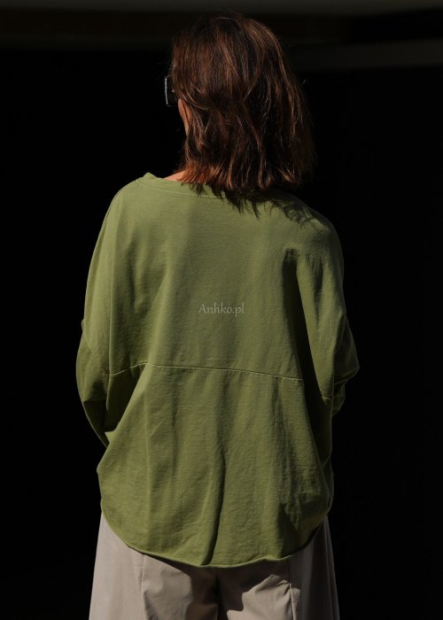 Zielona bluza oversize Nora z obniżoną linią ramion, długimi rękawami i dekoracyjną naszywką przy dole, zestawiona ze spodniami w podobnej tonacji.
