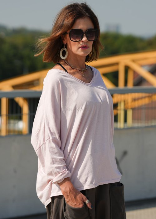 Pudroworóżowa bluzka damska oversize z długim rękawem i dekoltem V. Model pokazany w stylizacji z ciemną spódnicą i biżuterią, w miejskim otoczeniu.