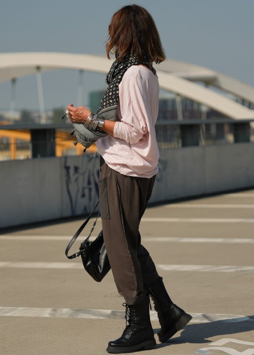 Pudroworóżowa bluzka damska oversize z długim rękawem i dekoltem V. Model pokazany w stylizacji z ciemną spódnicą i biżuterią, w miejskim otoczeniu.