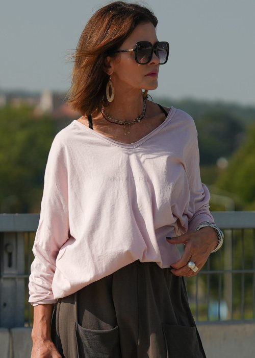 Pudroworóżowa bluzka damska oversize z długim rękawem i dekoltem V. Model pokazany w stylizacji z ciemną spódnicą i biżuterią, w miejskim otoczeniu.