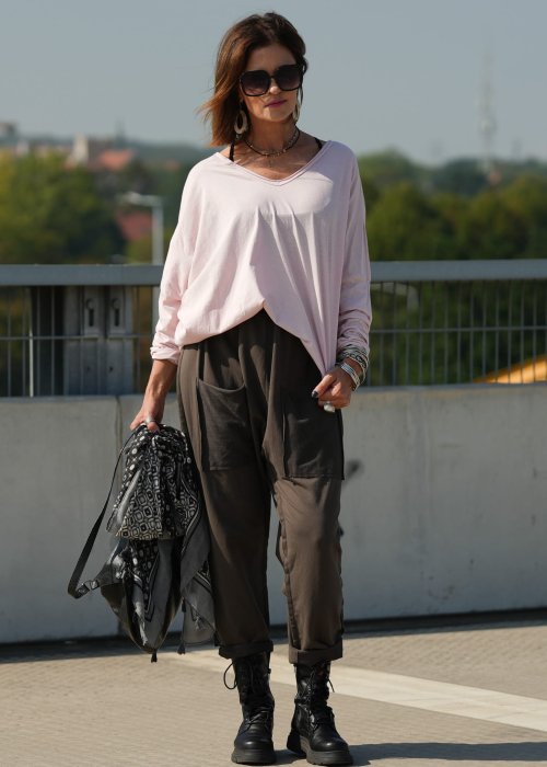 Pudroworóżowa bluzka damska oversize z długim rękawem i dekoltem V. Model pokazany w stylizacji z ciemną spódnicą i biżuterią, w miejskim otoczeniu.