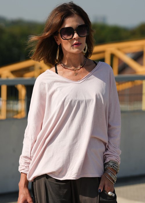 Pudroworóżowa bluzka damska oversize z długim rękawem i dekoltem V. Model pokazany w stylizacji z ciemną spódnicą i biżuterią, w miejskim otoczeniu.