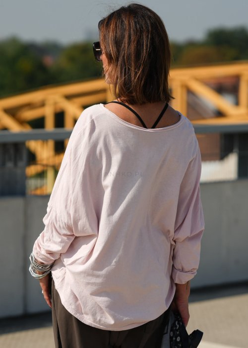 Pudroworóżowa bluzka damska oversize z długim rękawem i dekoltem V. Model pokazany w stylizacji z ciemną spódnicą i biżuterią, w miejskim otoczeniu.