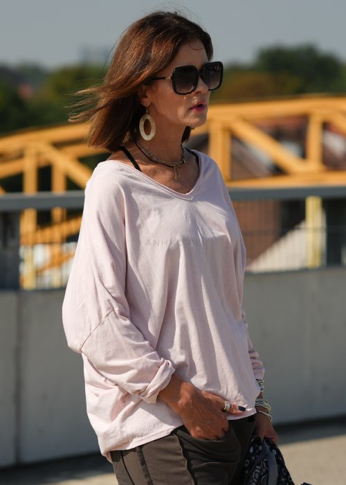 Pudroworóżowa bluzka damska oversize z długim rękawem i dekoltem V. Model pokazany w stylizacji z ciemną spódnicą i biżuterią, w miejskim otoczeniu.