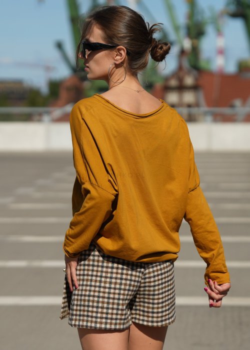 Musztardowa bluzka damska oversize z długim rękawem i dekoltem V wykończonym na surowo. Model pokazany w stylizacji z krótką spódnico-spodenką w kratę.