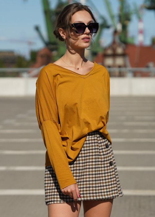Musztardowa bluzka damska oversize z długim rękawem i dekoltem V wykończonym na surowo. Model pokazany w stylizacji z krótką spódnico-spodenką w kratę.