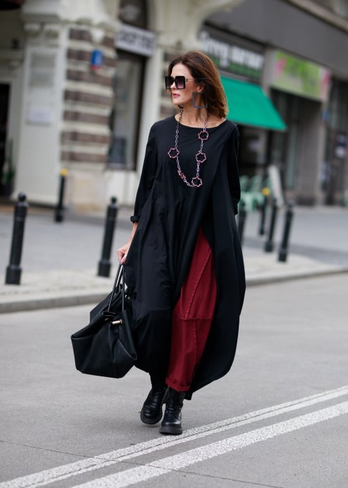 Czarna sukienka maxi o asymetrycznym dole i luźnym kroju oversize. Modelka prezentuje stylizację z czarnymi botkami, kapeluszem i minimalistyczną biżuterią w miejskim stylu.