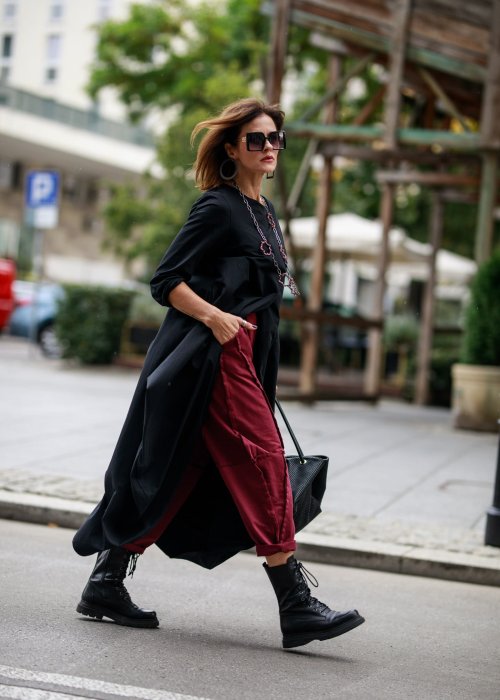 Czarna sukienka maxi o asymetrycznym dole i luźnym kroju oversize. Modelka prezentuje stylizację z czarnymi botkami, kapeluszem i minimalistyczną biżuterią w miejskim stylu.