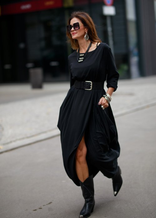 Czarna sukienka maxi o asymetrycznym dole i luźnym kroju oversize. Modelka prezentuje stylizację z czarnymi botkami, kapeluszem i minimalistyczną biżuterią w miejskim stylu.