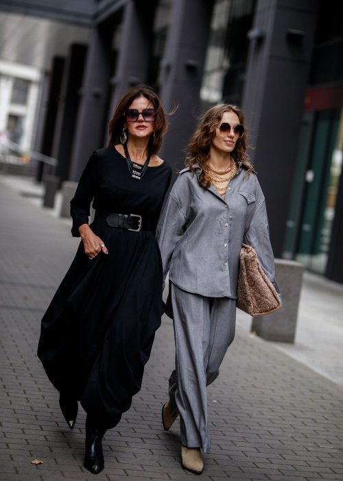 Czarna sukienka maxi o asymetrycznym dole i luźnym kroju oversize. Modelka prezentuje stylizację z czarnymi botkami, kapeluszem i minimalistyczną biżuterią w miejskim stylu.