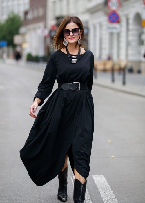 Czarna sukienka maxi o asymetrycznym dole i luźnym kroju oversize. Modelka prezentuje stylizację z czarnymi botkami, kapeluszem i minimalistyczną biżuterią w miejskim stylu.