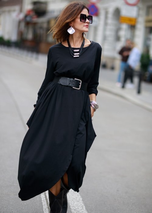 Czarna sukienka maxi o asymetrycznym dole i luźnym kroju oversize. Modelka prezentuje stylizację z czarnymi botkami, kapeluszem i minimalistyczną biżuterią w miejskim stylu.