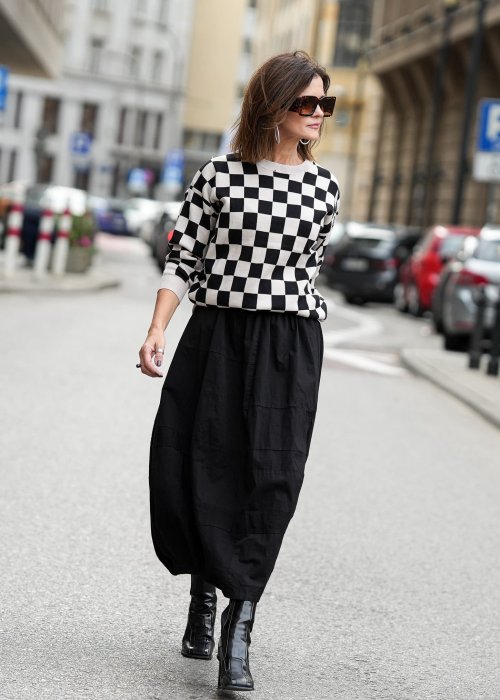 Czarna spódnica damska o kroju bombki z kieszeniami i dekoracyjnymi przeszyciami, pokazana w miejskiej stylizacji z czarno-białym swetrem w szachownicę i botkami na obcasie.