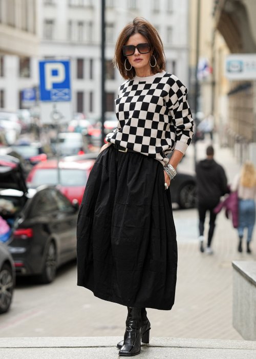 Czarna spódnica damska o kroju bombki z kieszeniami i dekoracyjnymi przeszyciami, pokazana w miejskiej stylizacji z czarno-białym swetrem w szachownicę i botkami na obcasie.