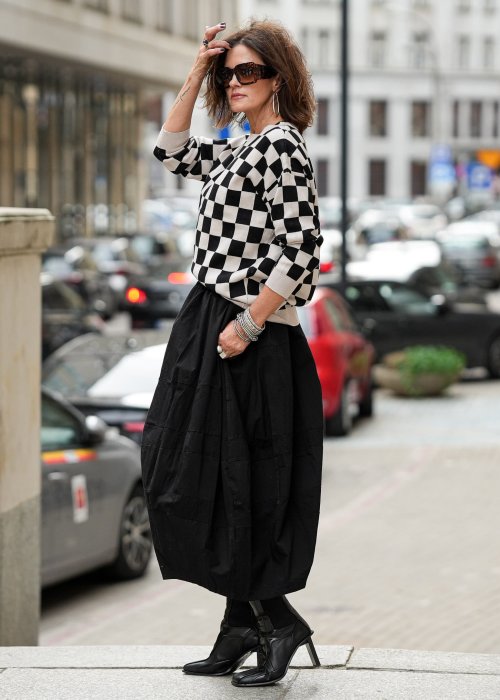 Czarna spódnica damska o kroju bombki z kieszeniami i dekoracyjnymi przeszyciami, pokazana w miejskiej stylizacji z czarno-białym swetrem w szachownicę i botkami na obcasie.