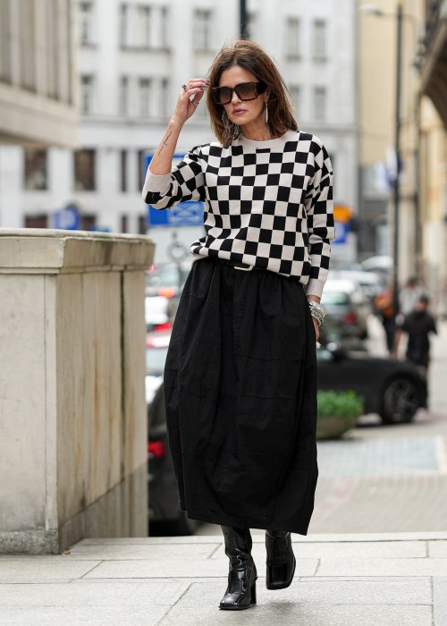 Czarna spódnica damska o kroju bombki z kieszeniami i dekoracyjnymi przeszyciami, pokazana w miejskiej stylizacji z czarno-białym swetrem w szachownicę i botkami na obcasie.