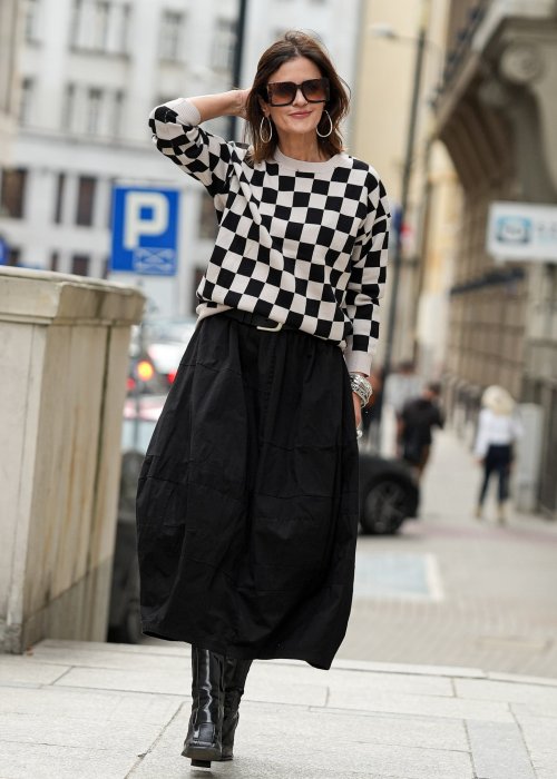 Czarna spódnica damska o kroju bombki z kieszeniami i dekoracyjnymi przeszyciami, pokazana w miejskiej stylizacji z czarno-białym swetrem w szachownicę i botkami na obcasie.
