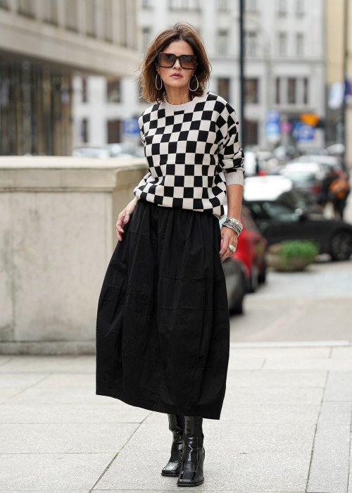 Czarna spódnica damska o kroju bombki z kieszeniami i dekoracyjnymi przeszyciami, pokazana w miejskiej stylizacji z czarno-białym swetrem w szachownicę i botkami na obcasie.