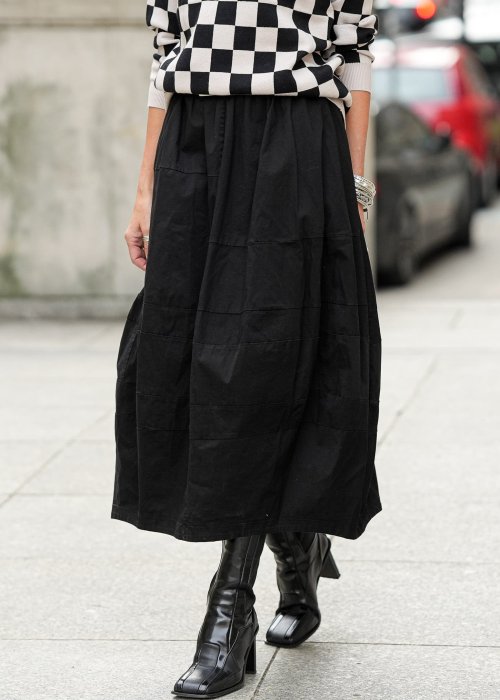 Czarna spódnica damska o kroju bombki z kieszeniami i dekoracyjnymi przeszyciami, pokazana w miejskiej stylizacji z czarno-białym swetrem w szachownicę i botkami na obcasie.