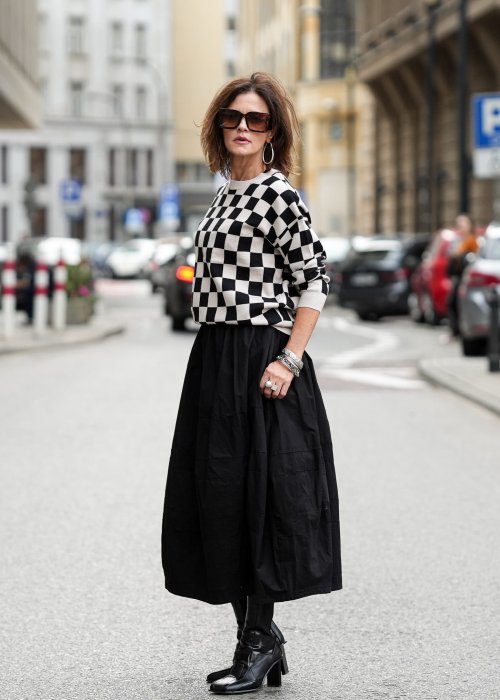 Czarna spódnica damska o kroju bombki z kieszeniami i dekoracyjnymi przeszyciami, pokazana w miejskiej stylizacji z czarno-białym swetrem w szachownicę i botkami na obcasie.