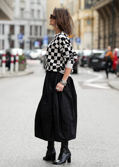 Czarna spódnica damska o kroju bombki z kieszeniami i dekoracyjnymi przeszyciami, pokazana w miejskiej stylizacji z czarno-białym swetrem w szachownicę i botkami na obcasie.