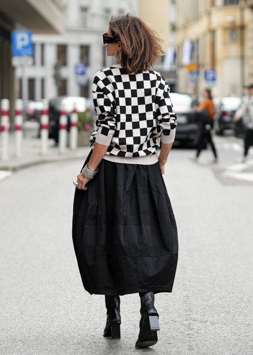 Czarna spódnica damska o kroju bombki z kieszeniami i dekoracyjnymi przeszyciami, pokazana w miejskiej stylizacji z czarno-białym swetrem w szachownicę i botkami na obcasie.