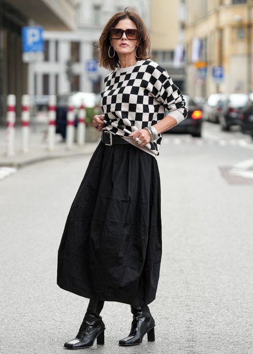Czarna spódnica damska o kroju bombki z kieszeniami i dekoracyjnymi przeszyciami, pokazana w miejskiej stylizacji z czarno-białym swetrem w szachownicę i botkami na obcasie.