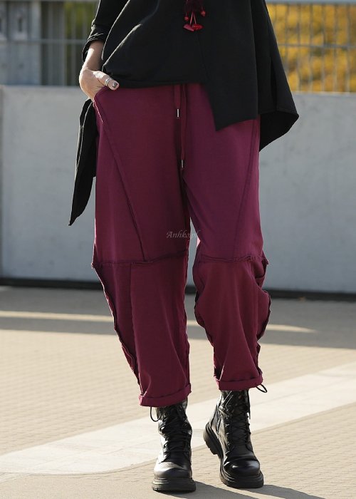 Bordowe spodnie dresowe damskie z obniżonym krokiem i ozdobnymi przeszyciami, luźny fason z gumką w pasie i sznurkiem, stylizacja miejska z czarnymi botkami.