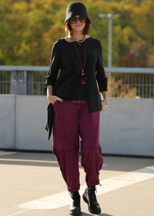 Bordowe spodnie dresowe damskie z obniżonym krokiem i ozdobnymi przeszyciami, luźny fason z gumką w pasie i sznurkiem, stylizacja miejska z czarnymi botkami.