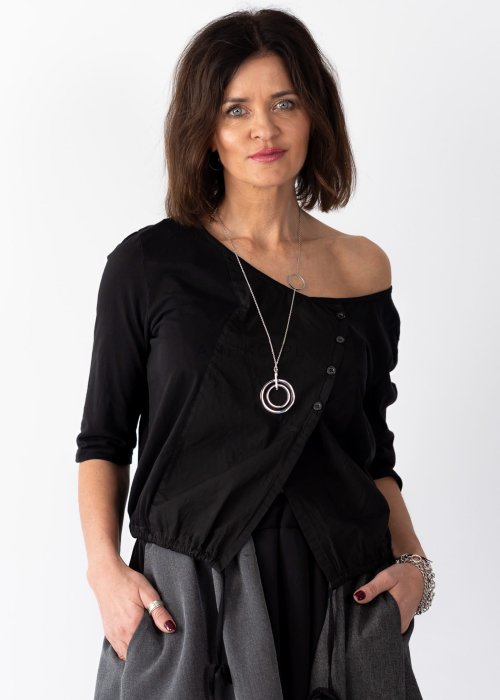 Czarna bluzka damska z asymetrycznym dekoltem odsłaniającym ramię i guzikami, stylizowana z elegancką spódnicą w minimalistycznym looku.