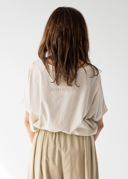 Kremowa bluzka damska oversize z szerokim dekoltem i krótkim rękawem w stylizacji z beżowymi spodniami i paskiem.
