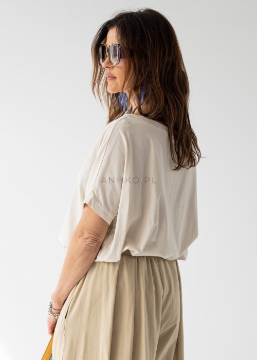 Kremowa bluzka damska oversize z szerokim dekoltem i krótkim rękawem w stylizacji z beżowymi spodniami i paskiem.