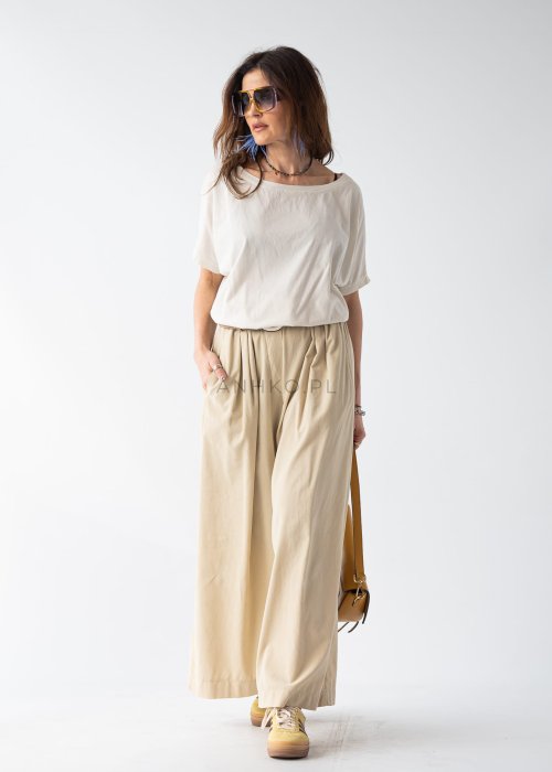 Kremowa bluzka damska oversize z szerokim dekoltem i krótkim rękawem w stylizacji z beżowymi spodniami i paskiem.