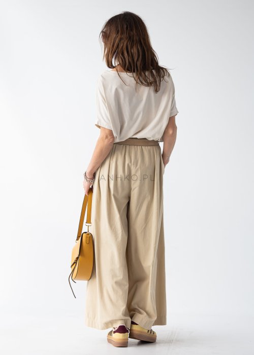 Kremowa bluzka damska oversize z szerokim dekoltem i krótkim rękawem w stylizacji z beżowymi spodniami i paskiem.
