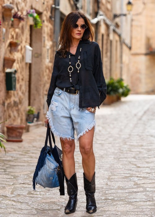 Jasne jeansowe spodenki damskie z przetarciami i asymetryczną, postrzępioną nogawką, stylizowane z czarną koszulą i paskiem w miejskim looku.