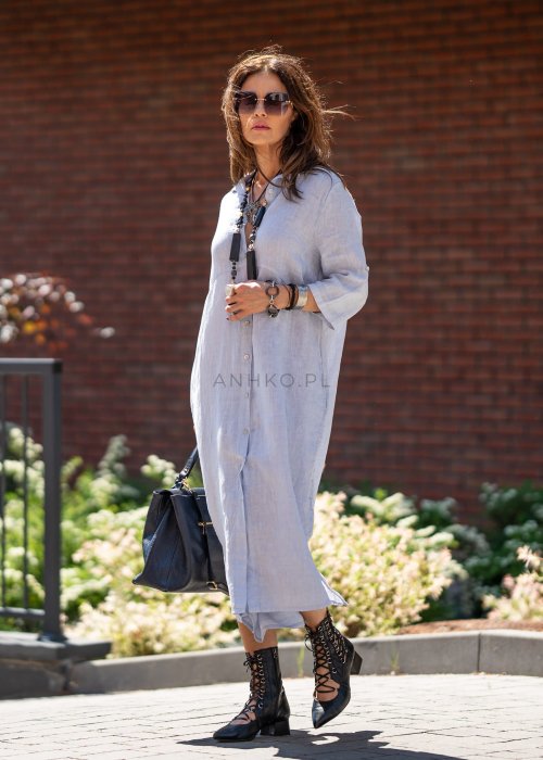 Jasnoniebieska lniana sukienka maxi w stylu szmizjerki, zapinana na guziki, z luźnym fasonem i kołnierzykiem, stylizowana z czarnymi botkami i torebką.