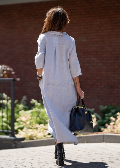 Jasnoniebieska lniana sukienka maxi w stylu szmizjerki, zapinana na guziki, z luźnym fasonem i kołnierzykiem, stylizowana z czarnymi botkami i torebką.