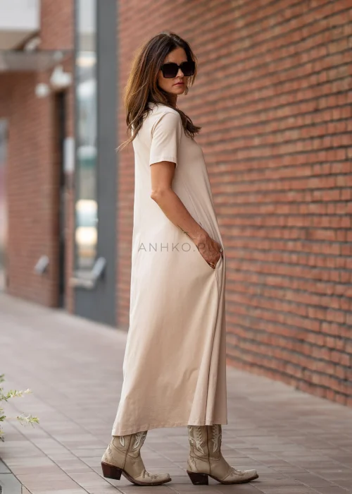 Beżowa sukienka maxi o luźnym kroju z krótkim rękawem i kieszeniami po bokach. Model pokazany w stylizacji z kowbojkami i złotą biżuterią.