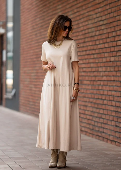 Beżowa sukienka maxi o luźnym kroju z krótkim rękawem i kieszeniami po bokach. Model pokazany w stylizacji z kowbojkami i złotą biżuterią.