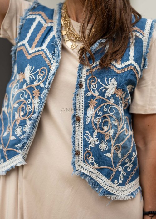 Niebieska kamizelka jeansowa damska z bogatymi haftami i surowym wykończeniem. Stylizacja z beżową sukienką i biżuterią w stylu boho.