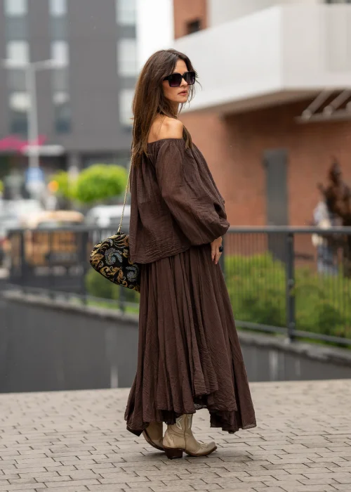 Brązowa bluzka hiszpanka oversize z gniecionej bawełny i szerokim dekoltem odsłaniającym ramiona. Stylizacja boho z długą spódnicą, dużą biżuterią i eleganckimi dodatkami.