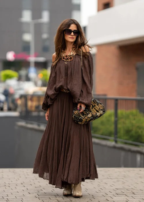 Brązowa bluzka hiszpanka oversize z gniecionej bawełny i szerokim dekoltem odsłaniającym ramiona. Stylizacja boho z długą spódnicą, dużą biżuterią i eleganckimi dodatkami.