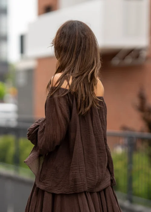Brązowa bluzka hiszpanka oversize z gniecionej bawełny i szerokim dekoltem odsłaniającym ramiona. Stylizacja boho z długą spódnicą, dużą biżuterią i eleganckimi dodatkami.
