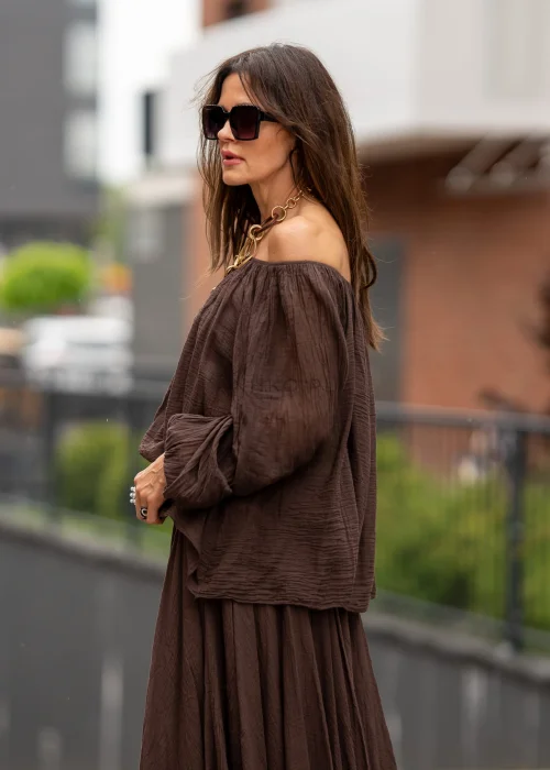 Brązowa bluzka hiszpanka oversize z gniecionej bawełny i szerokim dekoltem odsłaniającym ramiona. Stylizacja boho z długą spódnicą, dużą biżuterią i eleganckimi dodatkami.