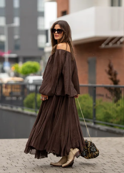 Brązowa bluzka hiszpanka oversize z gniecionej bawełny i szerokim dekoltem odsłaniającym ramiona. Stylizacja boho z długą spódnicą, dużą biżuterią i eleganckimi dodatkami.