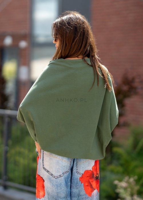Ciemnozielona bluzka oversize z szerokim rękawem typu nietoperz i opadającym dekoltem, zestawiona z jeansami z czerwonym printem w miejskiej stylizacji.