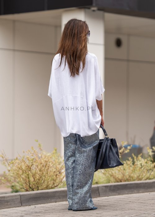 Biała bluzka oversize z lnem o szerokim kroju i krótkim rękawie, z dłuższym tyłem oraz subtelnym łączeniem materiałów. Model pokazany w stylizacji z połyskującymi spodniami i czarną torebką.